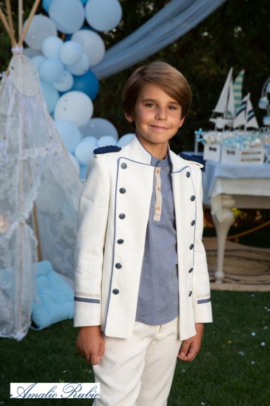 Comunión niño Niño vestido de traje blanco con detalles azules, sonríe en un ambiente festivo.